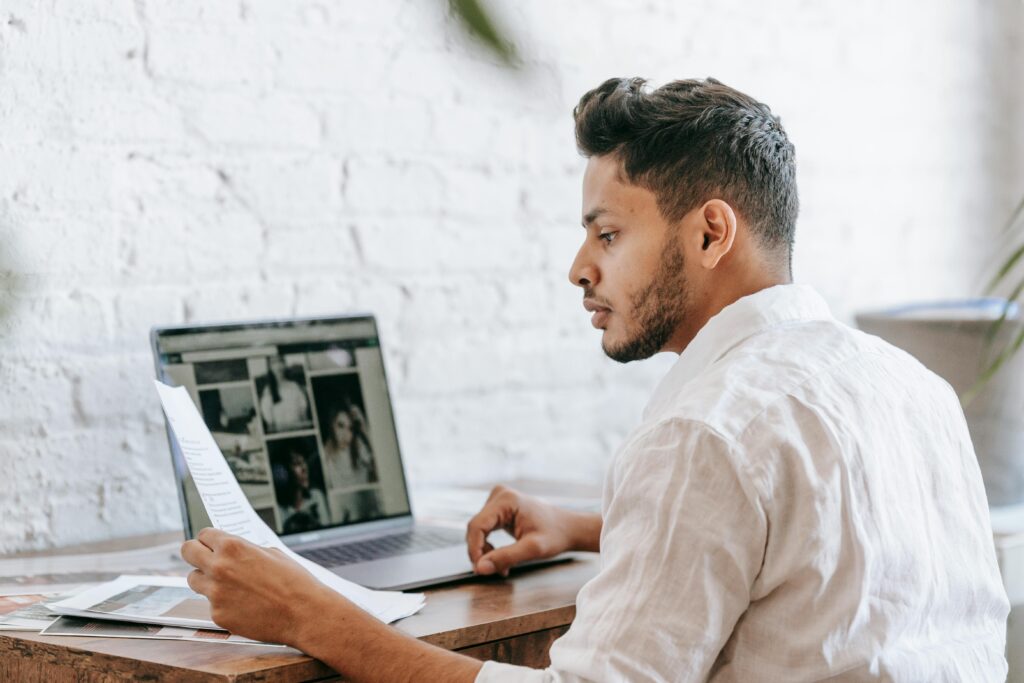 Back view focused ethnic male in white shirt reading document while sitting at desk with netbook in light office